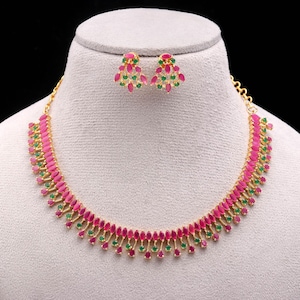 May include: A gold-toned necklace and matching earrings set. The jewelry features a repeating pattern of pink and green gemstones. The necklace and earrings are displayed on a mannequin head against a black background.
