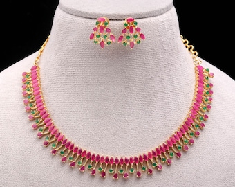 May include: A gold-toned necklace and matching earrings set. The jewelry features a repeating pattern of pink and green gemstones. The necklace and earrings are displayed on a mannequin head against a black background.