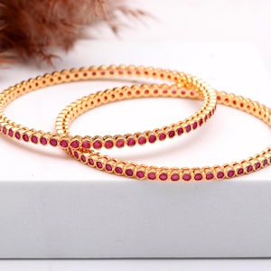 Peut inclure: Deux bracelets dorés ornés d'une rangée continue de petites pierres précieuses rouges rondes. Les bracelets sont présentés sur une surface blanche, avec un fond marron flou. Bijoux pour femmes.