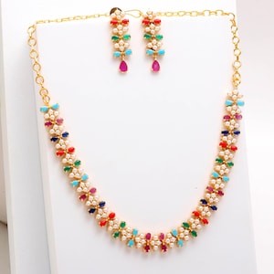 May include: A gold necklace and earring set. The necklace and earrings feature a floral design with colourful gemstones in red, green, blue, and purple, interspersed with small white pearls. The earrings have a teardrop-shaped pendant.
