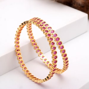 May include: Two gold bangles, each adorned with a repeating pattern of oval, magenta gemstones. The bangles are presented on a white surface, highlighting their intricate design and vibrant colour.