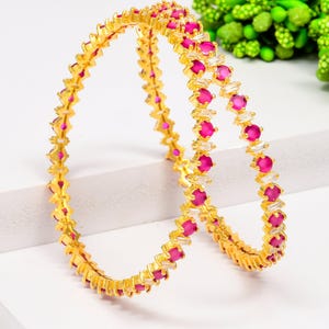 Könnte beinhalten: Zwei goldene Armreifen mit abwechselnd rosa und weißen Edelsteinen. Die Armreifen liegen auf einer weißen Oberfläche mit einem verschwommenen grünen Hintergrund. Die rosa Edelsteine sind rund, die weißen rechteckig.