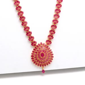 Peut inclure: Un collier doré orné d'un motif floral et serti de pierres précieuses roses vives. Le collier présente un pendentif central avec un grand groupe de pierres et une pierre en forme de goutte. La chaîne est ornée de plus petits groupes floraux.