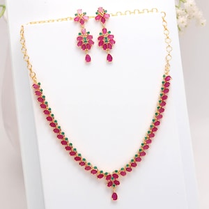 Op de afbeelding: Een goudkleurige sieradenset met een ketting en bijpassende oorbellen. De ketting en oorbellen zijn versierd met robijnrode en smaragdgroene edelstenen. De oorbellen hebben een bloemmotief met een druppelvormige edelsteen.