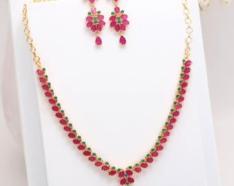 May include: A gold-toned jewelry set featuring a necklace and matching earrings. The necklace and earrings are adorned with ruby-red and emerald-green gemstones. The earrings have a floral design with a teardrop-shaped gemstone at the bottom.