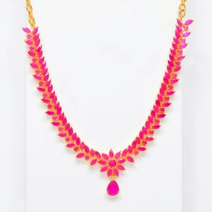 May include: A gold necklace with a striking pink gemstone design. The necklace features a floral pendant with a teardrop-shaped gemstone and a chain of pink gemstones. The necklace is set against a white background.
