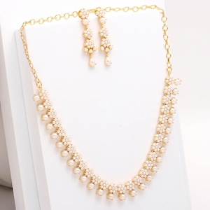 May include: A gold-toned necklace and earring set. The necklace and earrings feature a floral design with small, round, white pearls. The earrings are dangling, and the necklace is a choker style. The jewelry is displayed on a white surface.