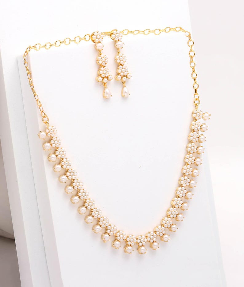 May include: A gold-toned necklace and earring set. The necklace and earrings feature a floral design with small, round, white pearls. The earrings are dangling, and the necklace is a choker style. The jewelry is displayed on a white surface.