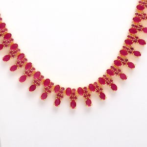 May include: A gold-toned necklace featuring a series of oval-shaped, ruby-red gemstones. The necklace has a delicate design with the gemstones set in a repeating pattern. The background is plain white, highlighting the jewelry.