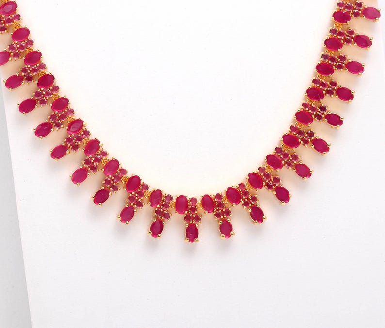 May include: A gold-toned necklace featuring a series of oval-shaped, ruby-red gemstones. The necklace has a delicate design with the gemstones set in a repeating pattern. The background is plain white, highlighting the jewelry.