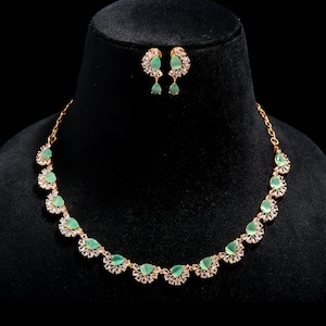 Puede incluir: Un conjunto de collar y pendientes de color dorado. El collar presenta piedras preciosas en forma de lágrima, de color verde claro, rodeadas de pequeños cristales transparentes. Los pendientes a juego tienen un diseño similar. Las joyas se exhiben en una cabeza de maniquí negra.