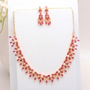 Puede incluir: Un conjunto de collar y pendientes dorados. El collar y los pendientes presentan piedras en forma de lágrima de color rubí y pequeñas perlas blancas. Las joyas se exhiben sobre una superficie blanca.