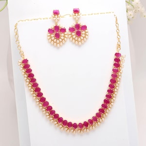 Könnte beinhalten: Ein goldfarbenes Schmuckset mit einer Halskette und passenden Ohrringen. Die Halskette und die Ohrringe sind mit ovalen, magentafarbenen Steinen und kleinen, weißen perlenartigen Akzenten verziert. Die Ohrringe haben ein Blumenmuster.