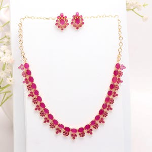 May include: A gold necklace and matching earrings set, featuring ruby-coloured oval gemstones. The necklace has a delicate chain. The jewellery is displayed on a white surface, with white flowers in the background.