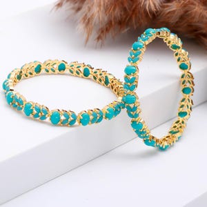 Peut inclure: Deux bracelets dorés ornés de pierres turquoise. Les bracelets, de forme ovale, présentent un motif répétitif de pierres turquoise serties dans du métal doré. Le fond est blanc.