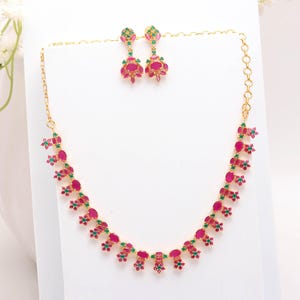 Puede incluir: Un conjunto de collar y pendientes dorados. El collar y los pendientes presentan un patrón repetido de piedras preciosas rojas y verdes. Los pendientes son colgantes y el collar es de estilo gargantilla.
