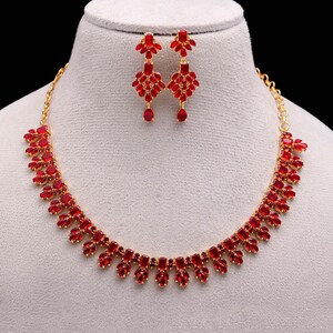 Puede incluir: Un conjunto de collar y pendientes a juego con piedras preciosas rojas. El collar y los pendientes tienen una base de metal dorado. El collar es de estilo gargantilla con piedras rojas. Los pendientes son de estilo colgante.