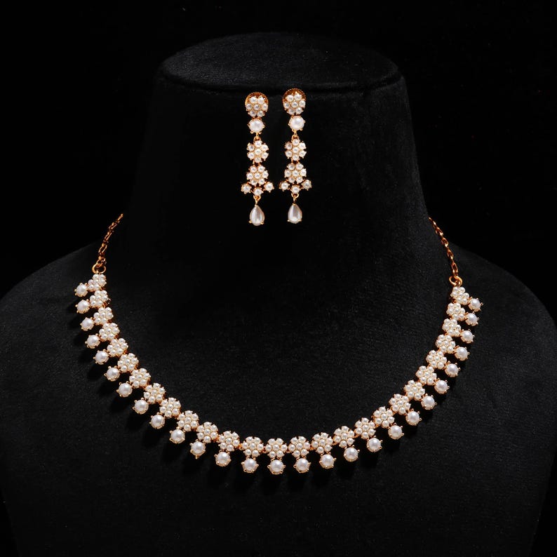 May include: A gold-toned necklace and earring set displayed on a black mannequin. The necklace and earrings feature a floral design with white pearl-like beads and clear, sparkling accents. The earrings dangle with a teardrop-shaped element.