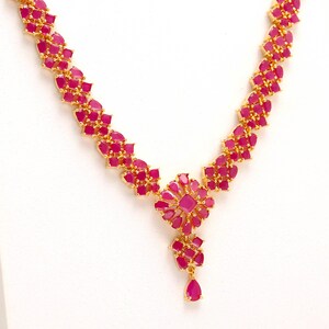 Pode incluir: Um colar dourado com um design floral, apresentando numerosas pedras preciosas rosa vibrantes. O colar tem um pingente central em forma de flor e um pingente em forma de gota. As pedras preciosas são definidas em um padrão repetitivo ao longo da corrente.