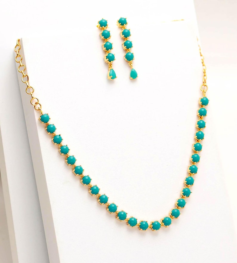 Op de afbeelding: Een goudkleurige ketting en bijpassende oorbellen set. De ketting en oorbellen zijn voorzien van turquoise, ronde edelstenen. De oorbellen hebben een druppelvormige edelsteen aan de onderkant. De sieraden worden gepresenteerd op een wit oppervlak.