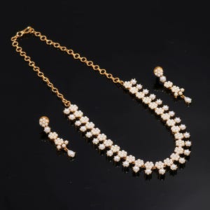 May include: A gold-toned necklace and earring set. The necklace and earrings feature a floral design with small, white pearls. The necklace has a chain closure. The earrings have a post closure with a dangling pearl.