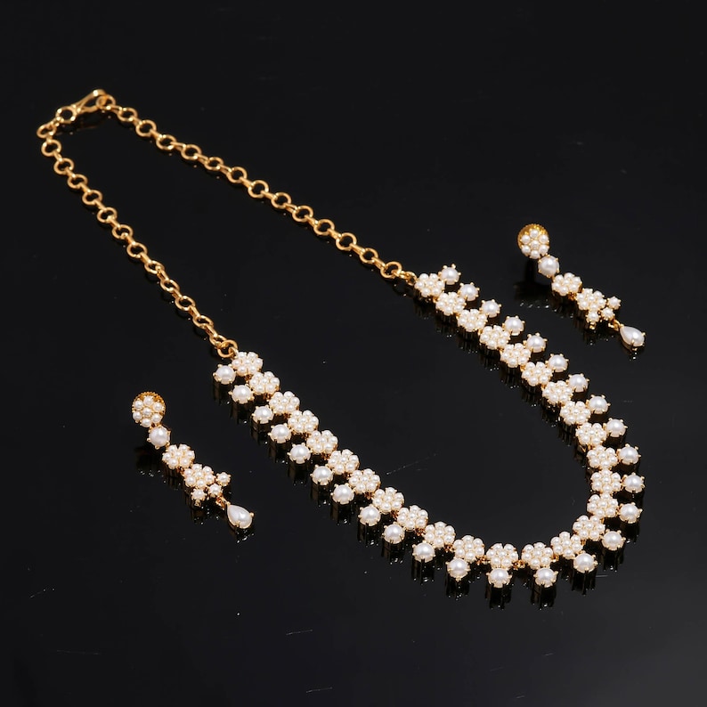 May include: A gold-toned necklace and earring set. The necklace and earrings feature a floral design with small, white pearls. The necklace has a chain closure. The earrings have a post closure with a dangling pearl.
