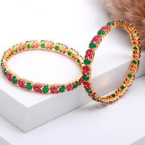 May include: Two gold bangles, each adorned with alternating green and red gemstones. The bangles are displayed on a white surface, highlighting their detailed design and vivid colours. The gemstones are arranged in a repeating pattern.