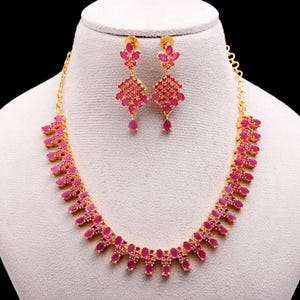 May include: A gold-toned necklace and earring set featuring vibrant pink gemstones. The necklace has a repeating pattern of oval and rectangular stones. The earrings have a floral design with dangling teardrop-shaped stones. The jewelry is displayed on a mannequin.