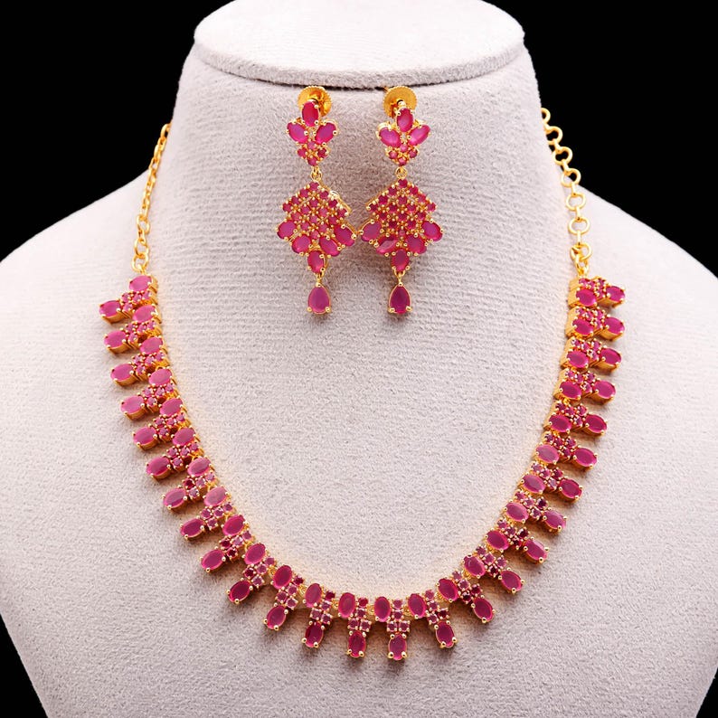 May include: A gold-toned necklace and earring set featuring vibrant pink gemstones. The necklace has a repeating pattern of oval and rectangular stones. The earrings have a floral design with dangling teardrop-shaped stones. The jewelry is displayed on a mannequin.
