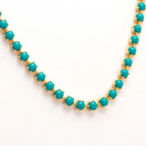Op de afbeelding: Een goudkleurige ketting met levendige turquoise kralen. Elke kraal is gezet in een delicate, goudkleurige zetting, waardoor een continue, elegante ketting ontstaat. De ketting wordt gepresenteerd tegen een witte achtergrond.