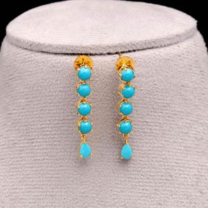 Op de afbeelding: Goudkleurige oorhangers met vijf turquoise kralen en een druppelvormige turquoise steen. De oorbellen worden gepresenteerd op een grijze ondergrond, die hun ingewikkelde ontwerp en levendige kleur benadrukt.