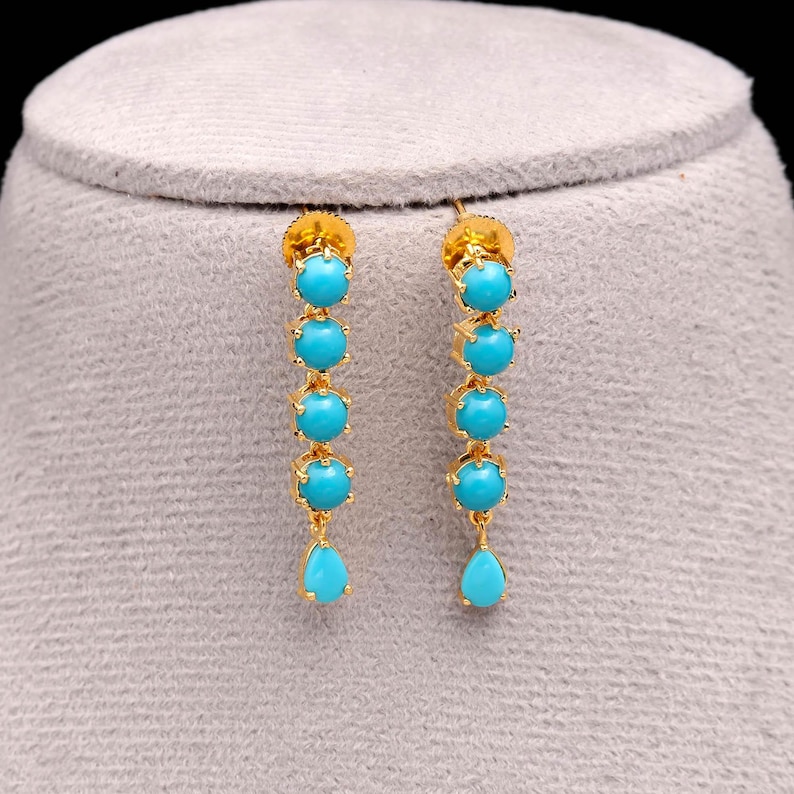 Op de afbeelding: Goudkleurige oorhangers met vijf turquoise kralen en een druppelvormige turquoise steen. De oorbellen worden gepresenteerd op een grijze ondergrond, die hun ingewikkelde ontwerp en levendige kleur benadrukt.