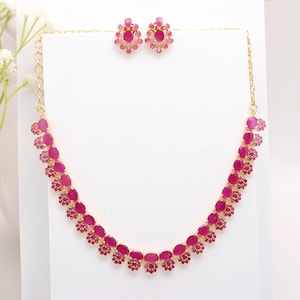 Puede incluir: Un conjunto de collar y pendientes a juego en tono dorado. El collar presenta una serie de piedras ovaladas de color magenta, cada una rodeada de pequeños detalles en rosa oscuro. Los pendientes tienen un diseño similar.