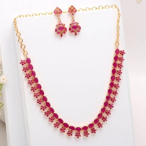 Puede incluir: Un conjunto de collar y pendientes a juego en tono dorado. El collar y los pendientes presentan piedras preciosas rosas oscuras de forma ovalada. Los pendientes son de estilo colgante y el collar tiene un diseño floral. Las joyas se exhiben sobre una superficie blanca.