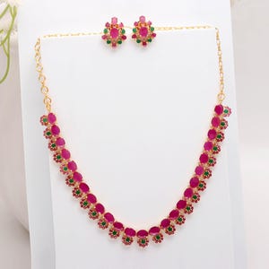 May include: A gold necklace and earring set. The necklace and earrings feature oval ruby-coloured stones with smaller green stones. The earrings are circular, and the necklace has a delicate chain. The set is displayed on a white surface.