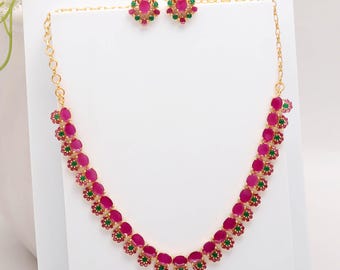 May include: A gold-toned necklace and matching earrings set. The necklace and earrings feature oval-shaped, ruby-colored stones interspersed with smaller green stones. The earrings are circular, and the necklace has a delicate chain.