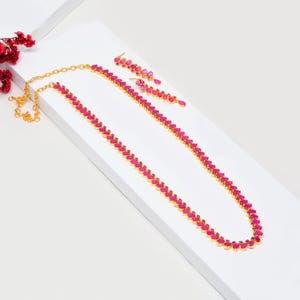 Puede incluir: Un collar dorado y pendientes a juego con vibrantes piedras preciosas rosas. El collar es largo y delicado, y los pendientes son de estilo colgante. Las joyas se exhiben sobre una superficie blanca, con flores rojas en el fondo.