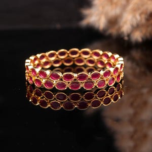 Può includere: Due braccialetti color oro con pietre rosso rubino di forma ovale. I braccialetti sono impilati, riflettendosi su una superficie scura. Le pietre preziose sono ravvicinate, creando un motivo continuo attorno ai braccialetti.