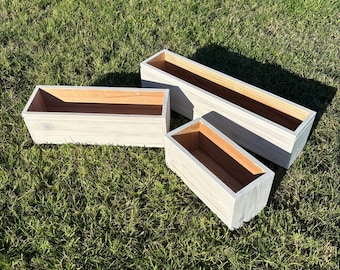 May include: Three rectangular wooden planters with a light gray finish and natural wood interiors are arranged on green grass. The planters vary in size, offering options for various plants and garden designs. These planters are ideal for outdoor or indoor use.