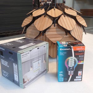 May include: Two boxes on a white surface. The box on the left is a light brown cardboard box with the words "METAL 3-LIGHT PENDANT KIT" printed on the front. The box on the right is a black cardboard box with the words "SMART BULB" printed on the front. A wooden light fixture is in the background.