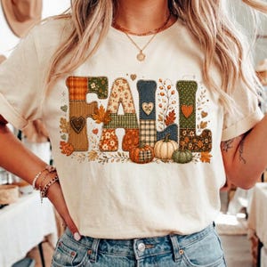 May include: A cream-coloured short-sleeve t-shirt featuring the word FALL in large, block-style letters. Each letter is filled with various patterns including plaid, floral, and polka dots in shades of orange, green, and brown, accented by pumpkins.