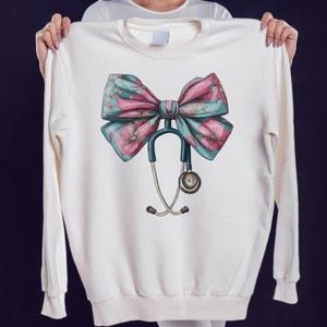 Può includere: Felpa bianca con un grande fiocco con fiori rosa e foglie turchesi e un design a stetoscopio. Il fiocco è il punto focale, con lo stetoscopio appeso sotto. Il design è centrato sulla parte anteriore della felpa.