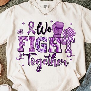 May include: Cream-colored t-shirt with a purple graphic that reads "We Fight Together." The design includes a purple ribbon, boxing gloves, and a flower. The text is in various fonts and patterns, including plaid and leopard print.