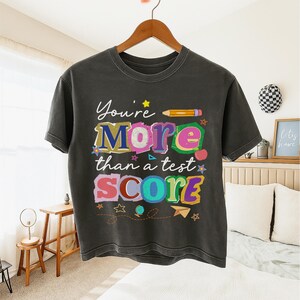 May include: Dark gray t-shirt with the text "You're More than a test SCORE" in colorful, playful lettering. The design includes a pencil, stars, and other decorative elements. The shirt is hanging on a wooden hanger.