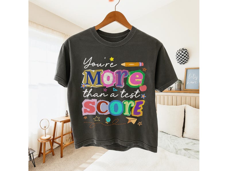 May include: Dark gray t-shirt with the text "You're More than a test SCORE" in colorful, playful lettering. The design includes a pencil, stars, and other decorative elements. The shirt is hanging on a wooden hanger.