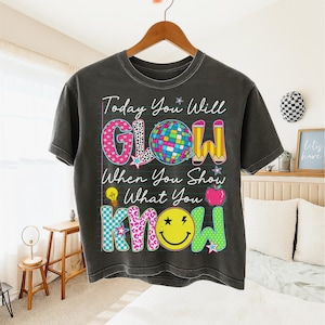 May include: Dark gray t-shirt with the text "Today You Will GLOW When You Show What You KNOW". The design includes a disco ball, pencils, a lightbulb, an apple, and a smiley face. The letters are in various patterns and colors.