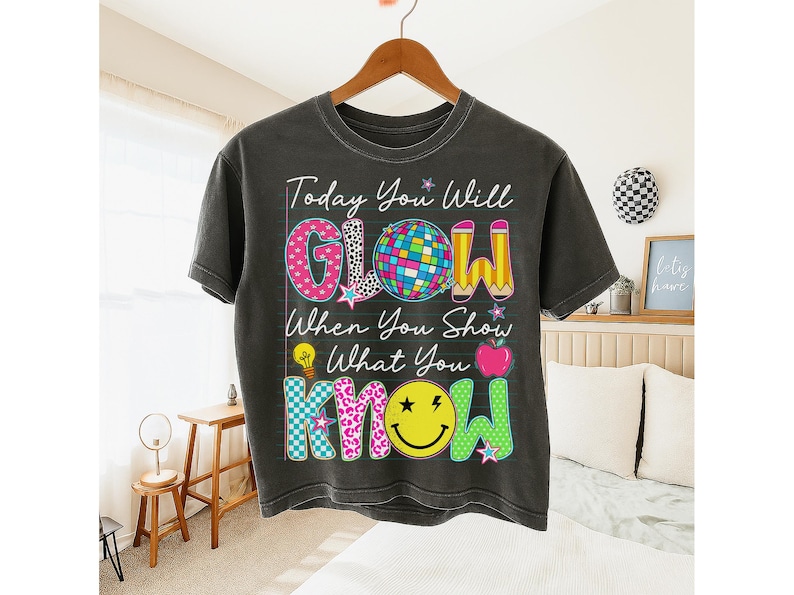 May include: Dark gray t-shirt with the text "Today You Will GLOW When You Show What You KNOW". The design includes a disco ball, pencils, a lightbulb, an apple, and a smiley face. The letters are in various patterns and colors.