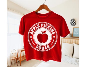 May include: Red t-shirt with a white circular graphic that reads "APPLE PICKING SQUAD" around a white apple illustration. The shirt is hanging on a wooden hanger.