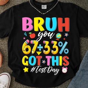 Op de afbeelding: Zwart T-shirt met een kleurrijk grafisch ontwerp. Het ontwerp bevat de woorden "BRUH you 67÷33% GOT THIS" in verschillende kleuren, samen met schoolthema-illustraties zoals potloden, een appel en een wereldbol. De tekst "#Test Day" is ook opgenomen.