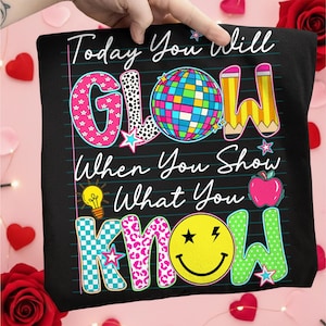 May include: Black t-shirt with the words "Today You Will GLOW When You Show What You KNOW" in colorful, playful lettering. The design includes a disco ball, pencils, a lightbulb, an apple, and a smiley face.